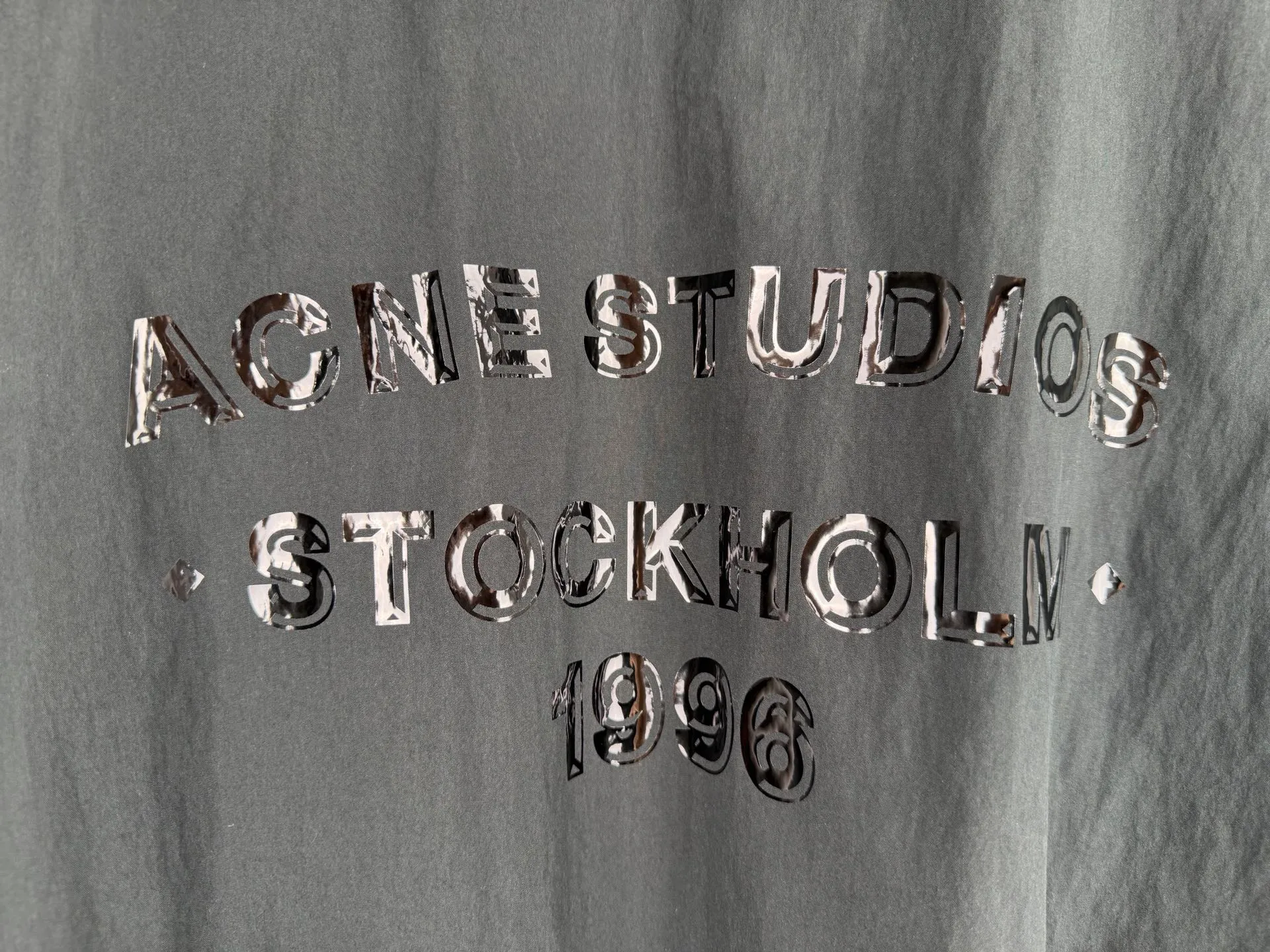 ACNE Studios back letter topstitch hooded jacket is made of fabric processing, contrasting topstitching, printing alignment and combination of elastic components. The fabric is treated with a physical wrinkle process, which does not rely on chemical coatings. It uses mechanical wrinkles to create a disordered texture effect on the surface of the fabric. While the fabric has a relaxed look, it still retains the structural tension of the woven fabric. During the cutting stage, the deformation caused by the folds of the fabric needs to be fully considered to ensure a stable silhouette after the pieces are spliced. The entire product uses contrasting topstitching as the main structural decoration. The seams of the cap, shoulder seams, cuffs, and hem are all exposed with contrasting topstitching. There are arc turns in different parts of the cut pieces. When sewing, it is necessary to control the fabric feeding rhythm and maintain a uniform stitch density. The elastic area of ​​the hem is sewn with multiple layers of materials. The layers of fabric, elastic bands, and hemming pieces are overlapped. It is easy for the line to shift and the fabric to arch, which requires high precision in the lathe operation. The Retro character printing on the back is the visual core of the entire garment. Multiple groups of characters are arranged in layers, giving it a worn and broken texture. The printing must ensure that the layer is strong and washable, but cannot cover up the details of the incomplete retro characters. At the same time, the printing positioning needs to be aligned with the middle seam of the back of the garment to avoid tilting and offset of the printing. The cuffs and hem are both elastic pleated structures. When sewing, you need to control the density of the fabric pleats to ensure a uniform and natural arc after shrinkage. The hood is spliced ​​into pieces and relies on multiple open threads to separate and shape it, ensuring that the hat is natural and three-dimensional and will not collapse against the face when worn. After all the sewing processes are completed, the garment needs to be ironed and finished to unify the wrinkles of the entire fabric and remove excess creases caused by the production process, finally presenting the complete Retro relaxed garment effect. Size: 46 48 50 52